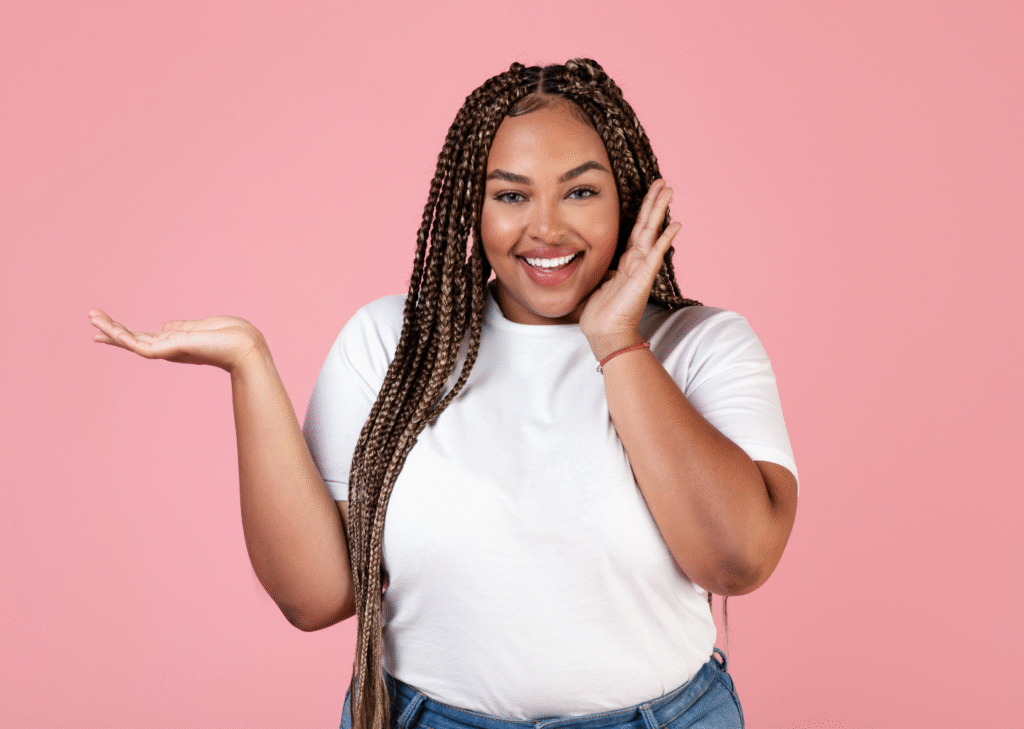 Smiling woman in a white t-shirt with braids, holding one hand up as if presenting something, standing in front of a pink background. Gastric balloon treatment representation.