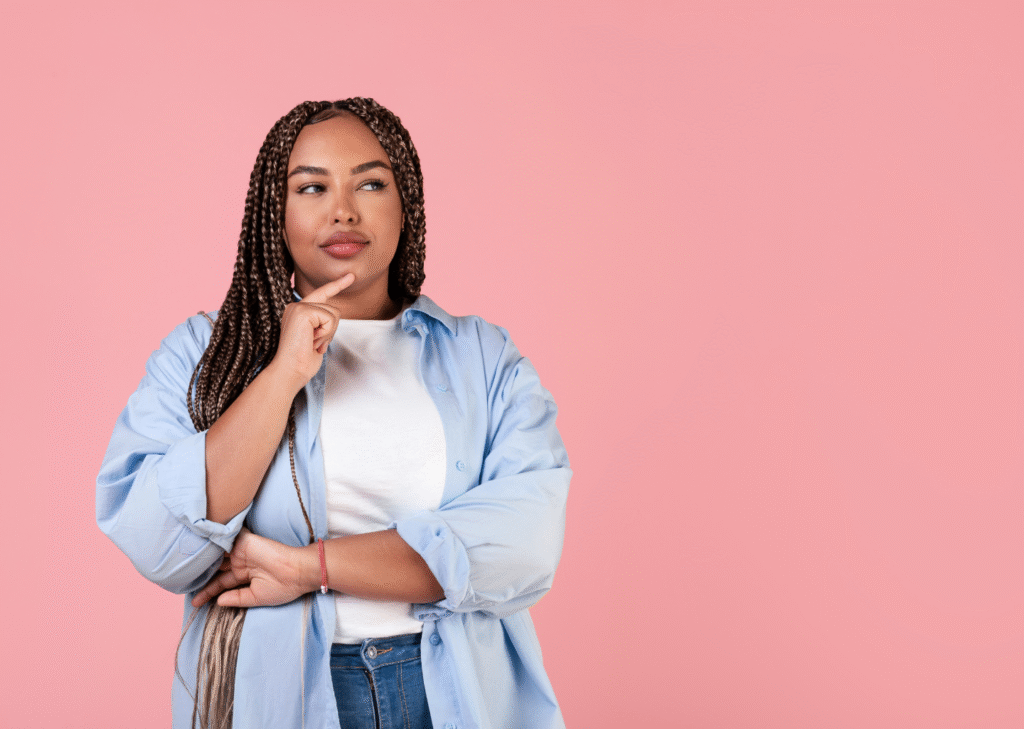 Thoughtful woman with braids wearing a blue shirt over a white t-shirt, posing with a hand on her chin in front of a pink background. Gastric balloon concept.