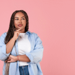 Thoughtful woman with braids wearing a blue shirt over a white t-shirt, posing with a hand on her chin in front of a pink background. Gastric balloon concept.