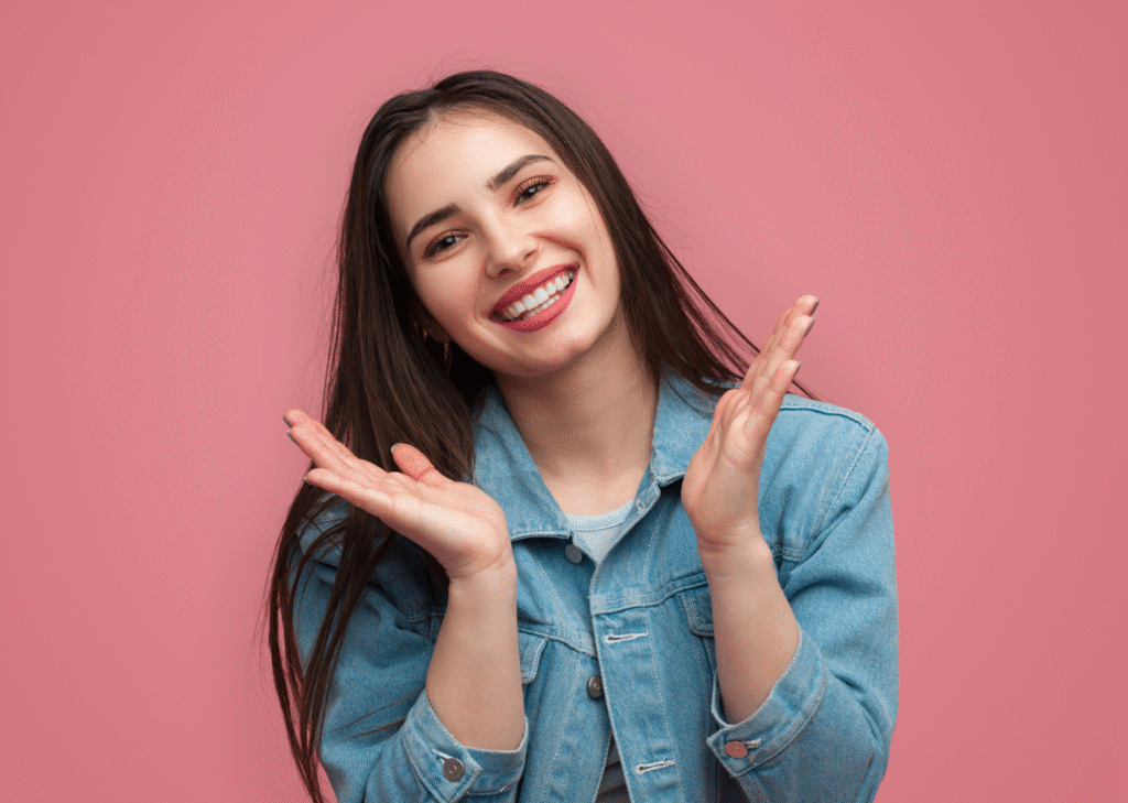 Smiling young woman after making hollywood smile with long brown hair wearing a denim jacket, posing cheerfully with hands raised near her face against a pink background.