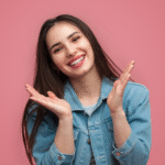 Smiling young woman after making hollywood smile with long brown hair wearing a denim jacket, posing cheerfully with hands raised near her face against a pink background.