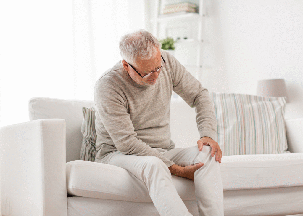 Elderly man sitting on a white couch, holding his knee in pain, possibly due to joint issues, and may be a candidate for Partial Knee Replacement.