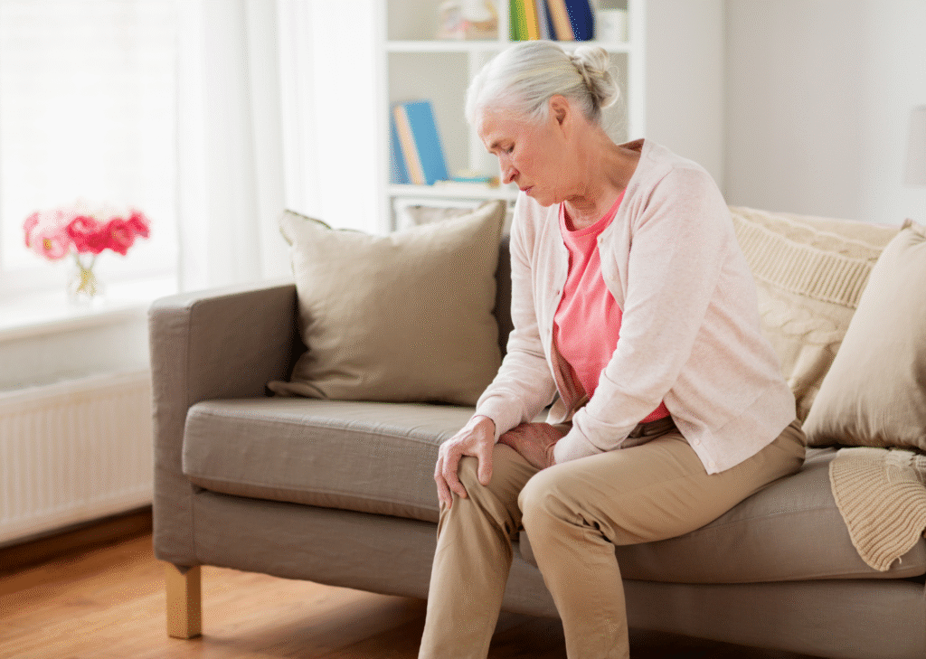 Elderly woman sitting on a beige sofa, grimacing in pain as she holds her knee, potentially indicating the need for a Partial Knee Replacement.