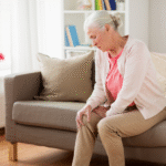 Elderly woman sitting on a beige sofa, grimacing in pain as she holds her knee, potentially indicating the need for a Partial Knee Replacement.