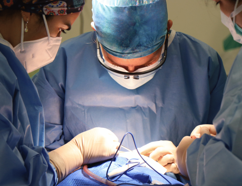 A close-up shot of three surgeons performing illeal interposition surgery. The central surgeon, wearing loupes, is focused on the surgical field, where gloved hands manipulate instruments and a wire or tube on blue surgical drapes. Two other surgeons, partially visible, assist in the procedure.