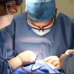 A close-up shot of three surgeons performing illeal interposition surgery. The central surgeon, wearing loupes, is focused on the surgical field, where gloved hands manipulate instruments and a wire or tube on blue surgical drapes. Two other surgeons, partially visible, assist in the procedure.