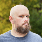 shorter A bald man with blue eyes and a full brown beard, possibly indicating a beard transplant, looks seriously at the camera against a blurred green and yellow background.