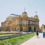 A wide shot captures the grand, ornate yellow building of the Croatian National Theatre in Zagreb under a clear, bright sky, a landmark often seen by those considering dental crowns in Zagreb.