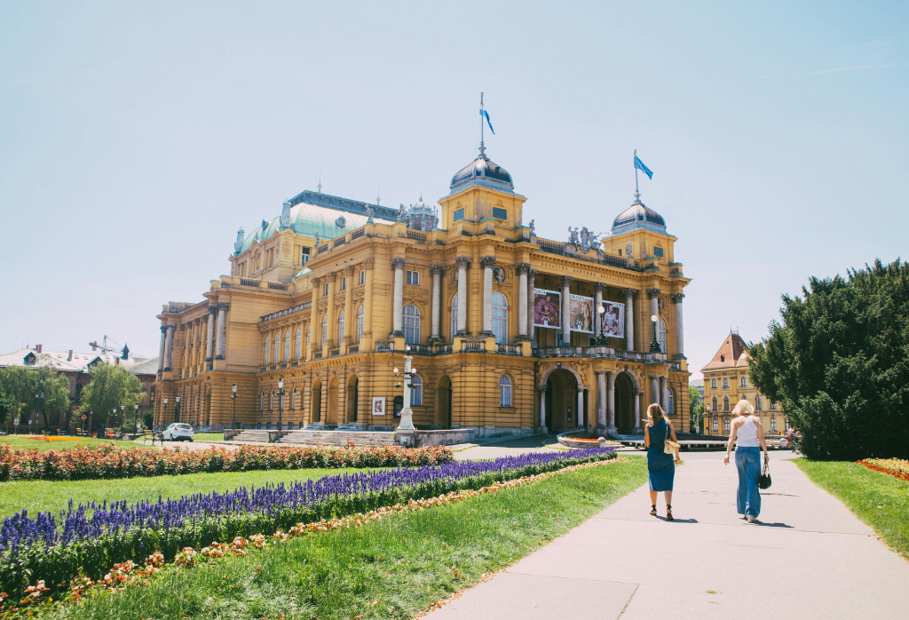 A wide shot captures the grand, ornate yellow building of the Croatian National Theatre in Zagreb under a clear, bright sky, a landmark often seen by those considering dental crowns in Zagreb.