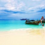 A serene tropical beach scene, ideal for plastic surgery holidays Thailand, features crystal-clear turquoise waters gently lapping against a pristine white sand shore under a partly cloudy blue sky.
