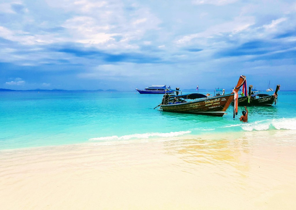 A serene tropical beach scene, ideal for plastic surgery holidays Thailand, features crystal-clear turquoise waters gently lapping against a pristine white sand shore under a partly cloudy blue sky.
