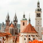 A scenic view of Prague's Old Town, featuring several historic buildings with distinctive red-tiled roofs and tall spires against a bright sky, often visited by those considering plastic surgery Prague.