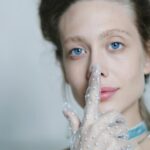 A fair-skinned woman with striking blue eyes gently touches her nose with a finger covered by a sheer, polka-dotted white glove, subtly hinting at the delicate nature of the rhinoplasty recovery time.