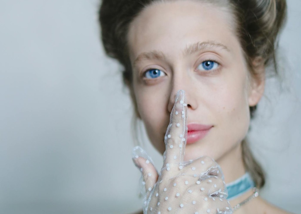 A fair-skinned woman with striking blue eyes gently touches her nose with a finger covered by a sheer, polka-dotted white glove, subtly hinting at the delicate nature of the rhinoplasty recovery time.