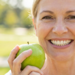Smiling woman with dental implants holding a green apple outdoors.
